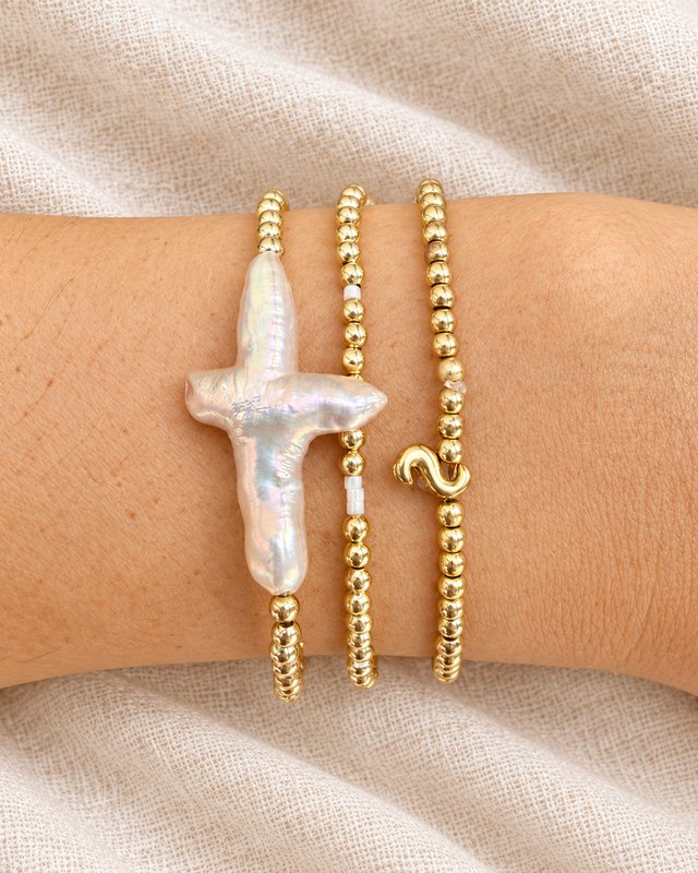 Three gold bead bracelets on a wrist, one with a baroque pearl cross and one with a gold S initial