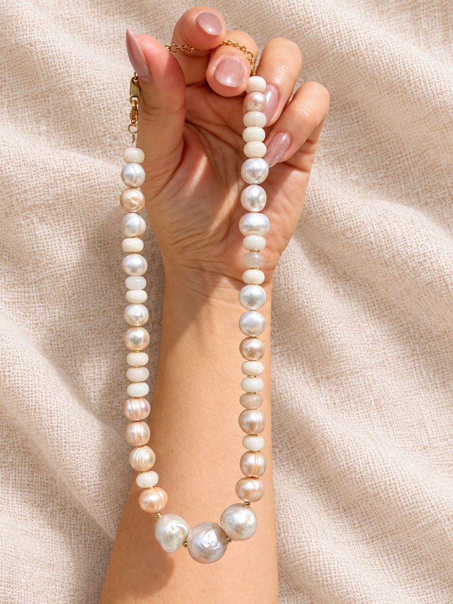 A freshwater pearl necklace in white and blush with larger baroque pearls at the center, held up in one hand against cream linen