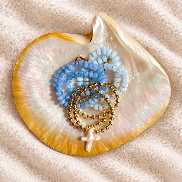 A mother-of-pearl shell dish holding blue gemstone necklaces, gold bead bracelets and a freshwater pearl cross