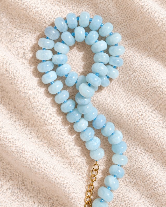 A light aqua gemstone necklace with a gold clasp laid in a loop on cream linen