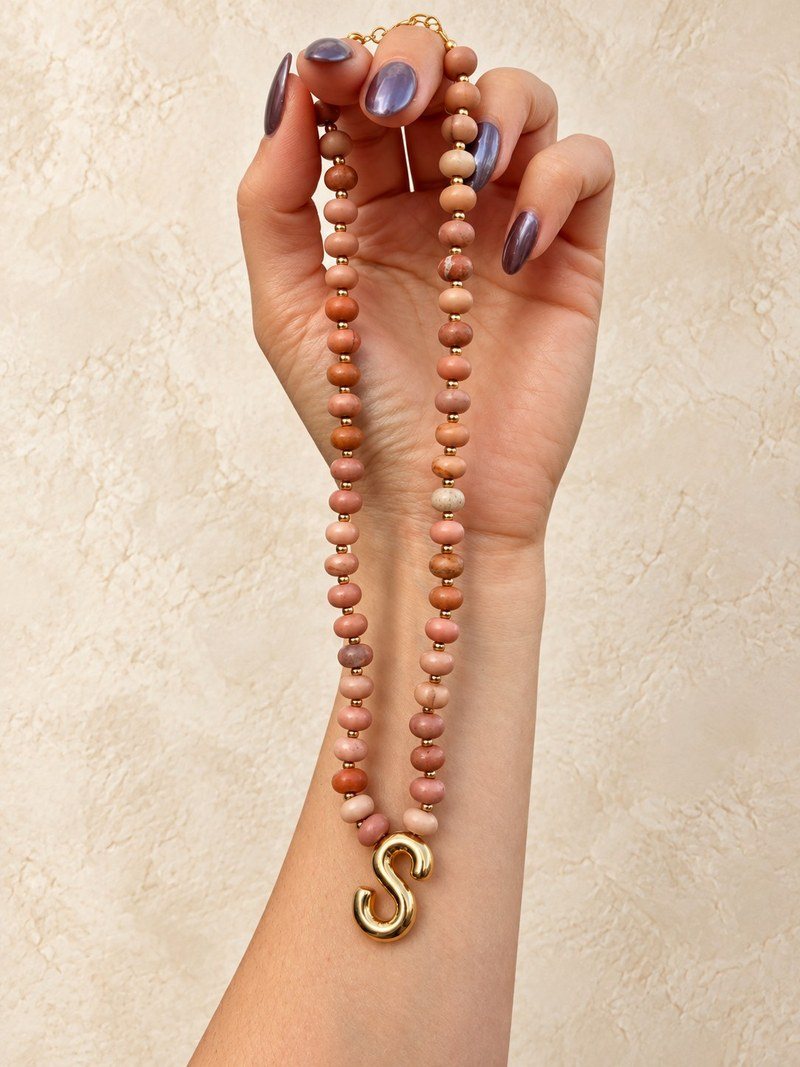 A blush semi-precious gemstone necklace with a polished gold S initial, held up in one hand