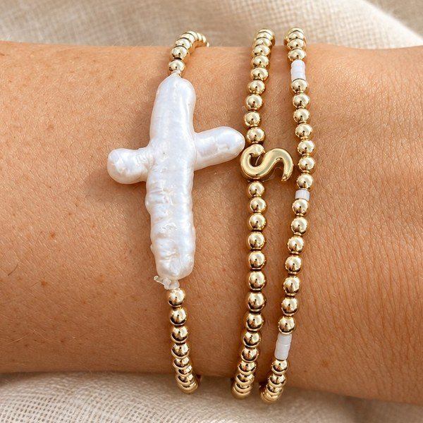 A stack of gold bead bracelets with a baroque pearl cross and a gold S initial