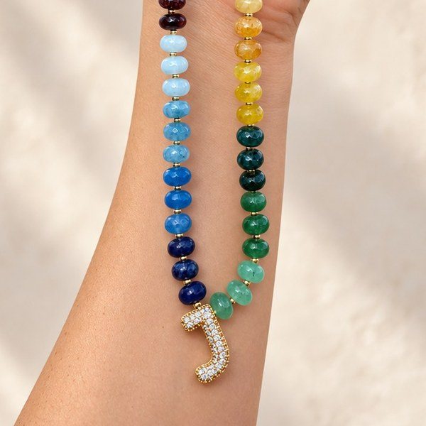 A rainbow semi-precious gemstone necklace with a pavé J initial, held up in one hand
